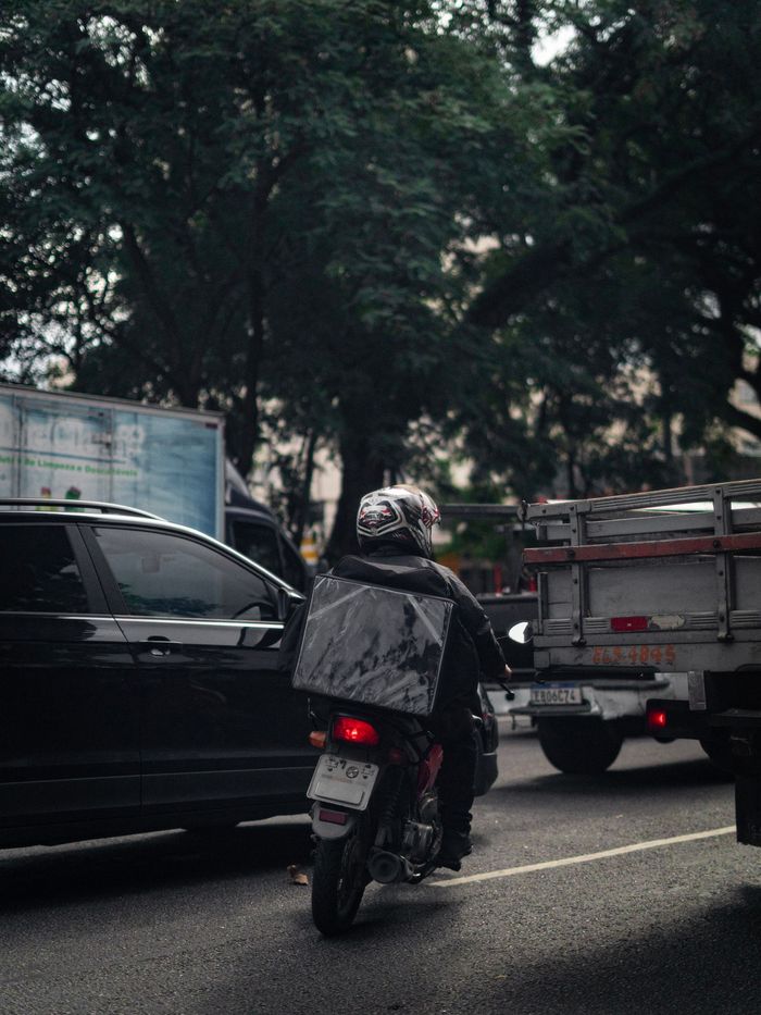 Motociclista visto por trás, entre um carro e um caminhão, no trânsito de uma avenida.